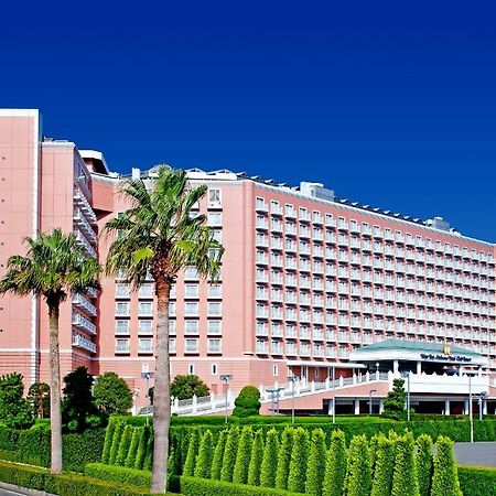 Grand Nikko Tokyo Bay Maihama Urayasu Exterior foto The image shows a large, multi-story hotel building featuring a light pink facade. The architecture appears modern, with numerous windows arranged in a grid pattern. In the foreground, there are neatly trimmed shrubs and palm trees, adding a tropical