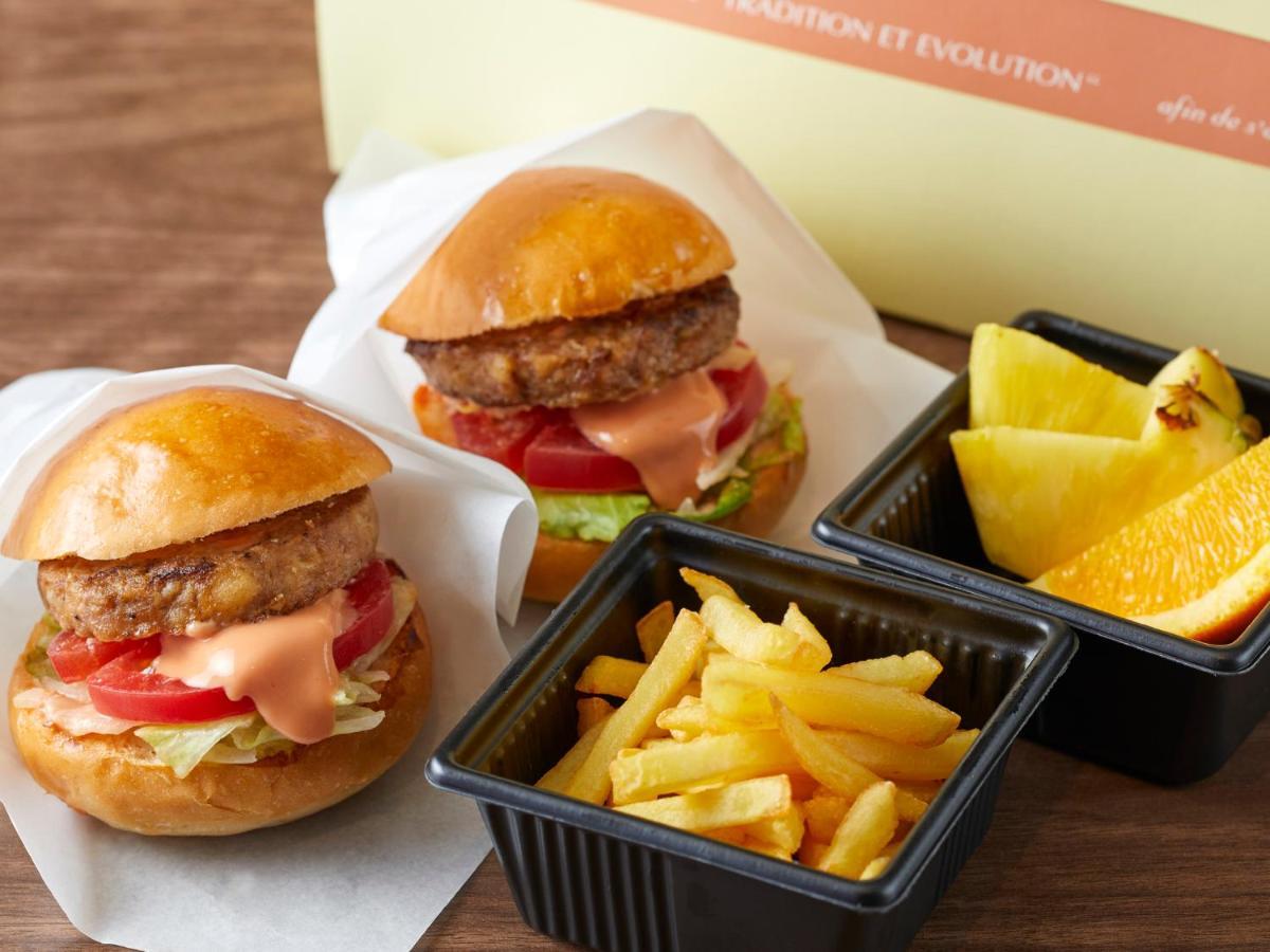 Grand Nikko Tokyo Bay Maihama Urayasu Exterior foto The photo shows two small burgers placed on a white background, each with a golden-brown bun, a fried patty, lettuce, tomato, and a creamy sauce. Alongside the burgers, there are two containers: one holding a serving of french fries and the other con