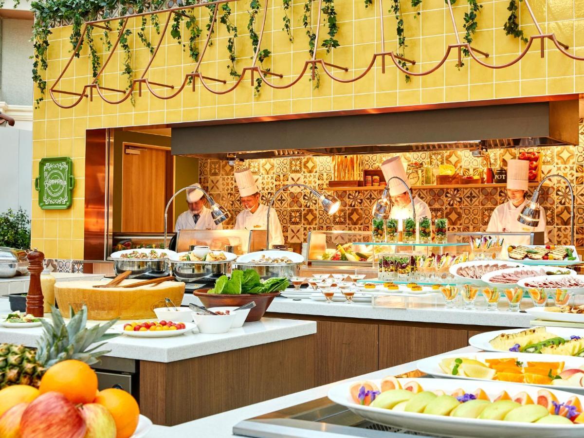 Grand Nikko Tokyo Bay Maihama Urayasu Exterior foto The photo shows a vibrant and colorful buffet setup in a restaurant. In the foreground, there are various dishes arranged on white platters, featuring an assortment of fruits, desserts, and other items. The background features chefs in white uniforms