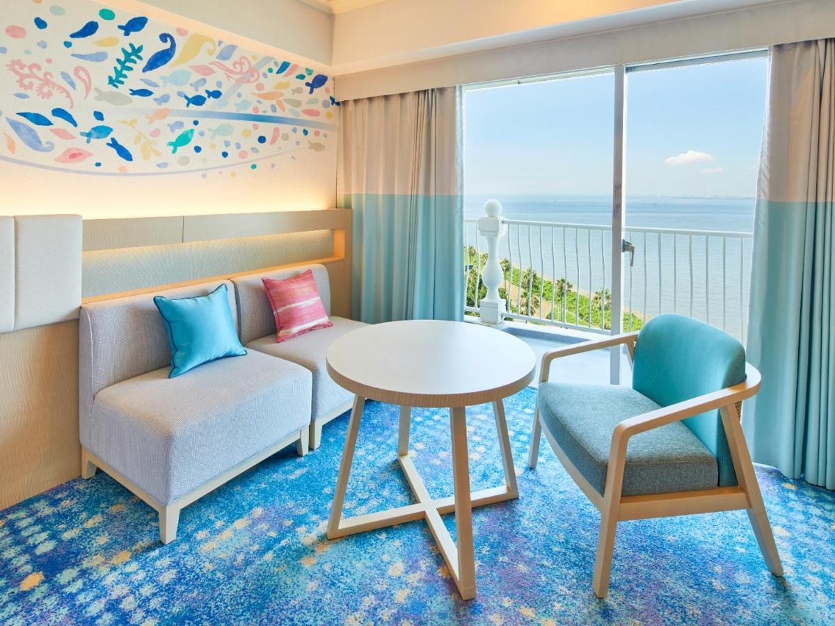 Grand Nikko Tokyo Bay Maihama Urayasu Exterior foto The photo shows a modern and stylish interior of a room with a view. There is a cozy seating area featuring a light-colored sofa with decorative cushions in various shades of blue and pink. A round table occupies the center of the space, surrounded b