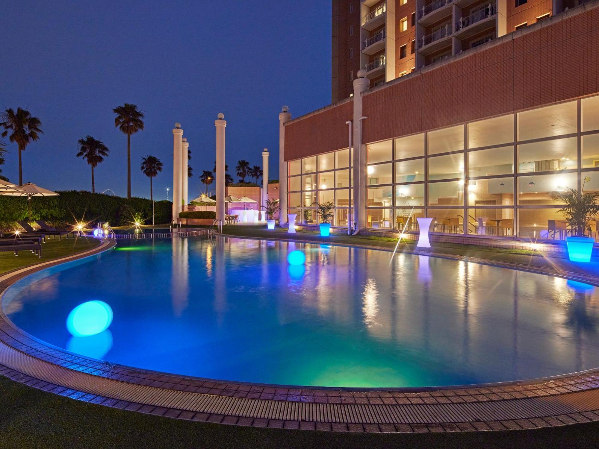 Grand Nikko Tokyo Bay Maihama Urayasu Exterior foto The photo shows a well-lit swimming pool at night, surrounded by a lush green area. The pool features glowing blue lights that create a serene and inviting atmosphere. Palm trees can be seen in the background, adding a tropical feel to the scene. Add