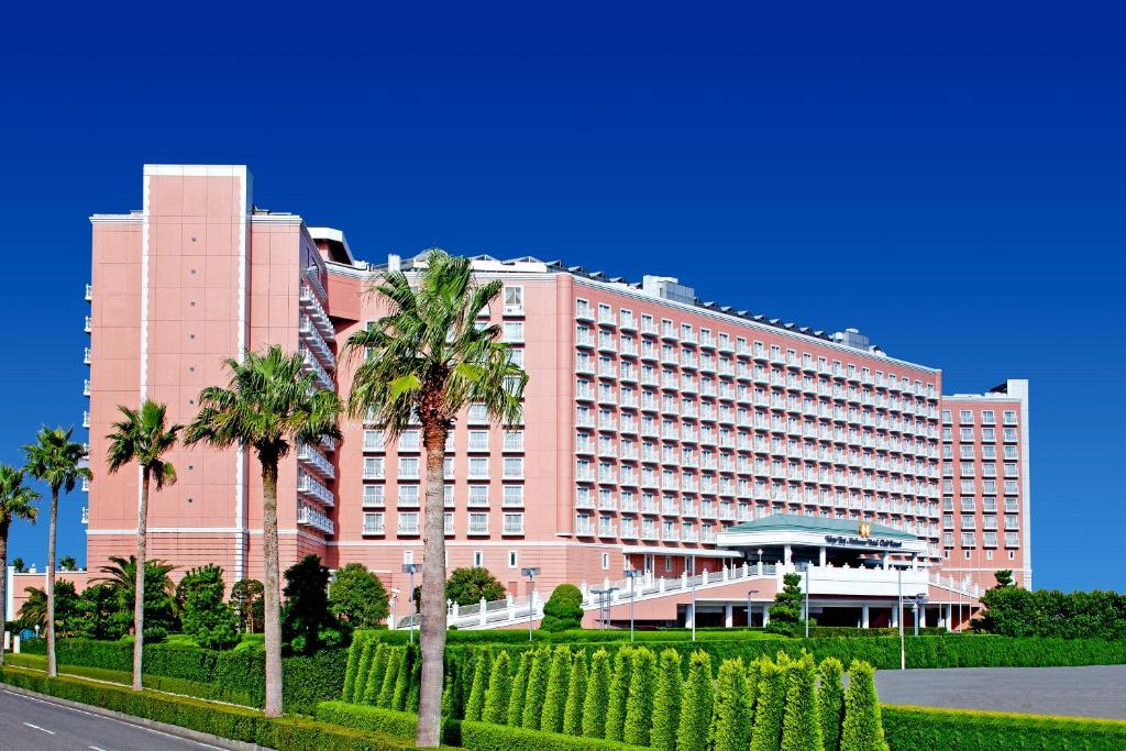 Grand Nikko Tokyo Bay Maihama Urayasu Exterior foto The image shows a large, multi-story hotel building featuring a light pink facade. The architecture appears modern, with numerous windows arranged in a grid pattern. In the foreground, there are neatly trimmed shrubs and palm trees, adding a tropical