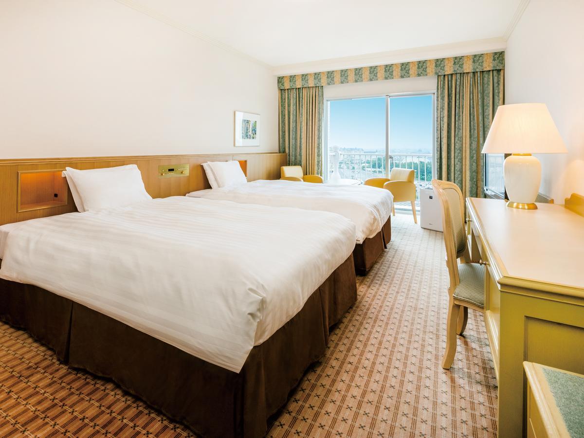 Grand Nikko Tokyo Bay Maihama Urayasu Exterior foto The photo shows a hotel room featuring two twin beds with white linens and dark brown bed skirts. The room has a light, neutral-colored wall and carpet. There is a window with curtains that let in natural light, offering a view outside. A small desk 