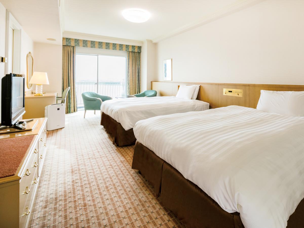 Grand Nikko Tokyo Bay Maihama Urayasu Exterior foto The photo shows a hotel room featuring two large beds with white bedding and brown bases. There's a light-colored carpet on the floor, and the room is brightly lit. To one side, there is a television on a stand and a small white refrigerator. A desk 